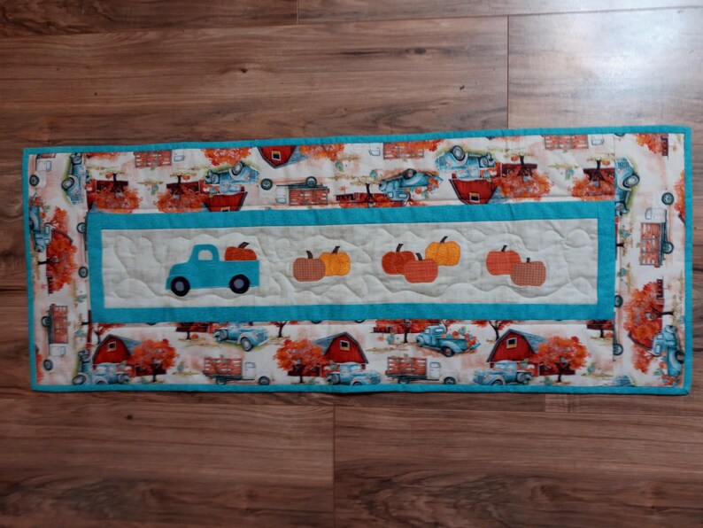 May include: A rectangular quilted table runner with a fall harvest theme. The runner features a central panel with a blue truck carrying pumpkins and several pumpkin appliqu&eacute;s. The border has a repeating pattern of trucks, barns, and fall foliage.