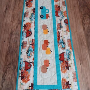 May include: A quilted table runner with a fall theme. The runner features a repeating pattern of blue trucks, red barns, and orange trees. A central panel displays a blue truck and several pumpkin appliqu&eacute;s, all bordered by a turquoise trim.