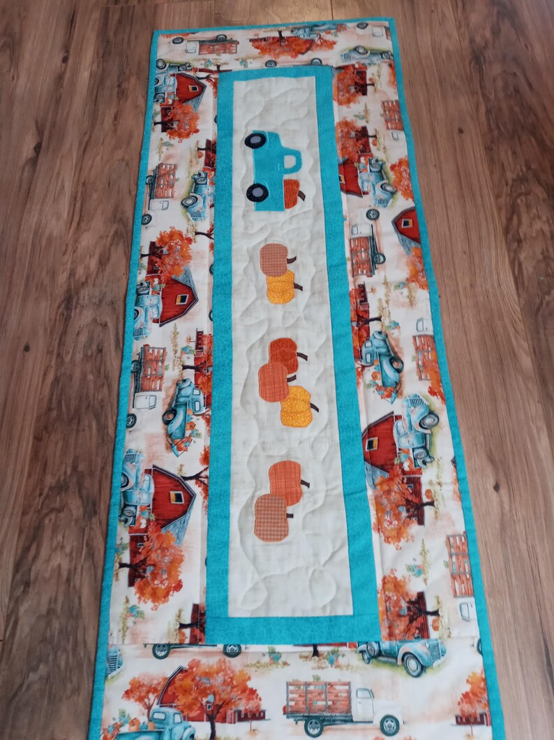 May include: A quilted table runner with a fall theme. The runner features a repeating pattern of blue trucks, red barns, and orange trees. A central panel displays a blue truck and several pumpkin appliqu&eacute;s, all bordered by a turquoise trim.