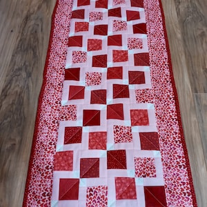 May include: A rectangular quilted table runner in shades of red and pink. The center features a pattern of alternating red squares, while the border is adorned with a heart-shaped pattern. The runner is approximately 60 inches long.