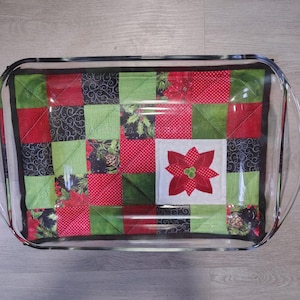 May include: A clear glass rectangular baking dish containing a quilted fabric hot pad. The hot pad features a patchwork design of red, green, and black fabric squares with patterns of poinsettias, pinecones, and dots, plus a central poinsettia.