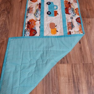 May include: A quilted table runner featuring a central panel with a blue truck and hay bales applique. The runner has a white background with a teal border and backing. The sides display a repeating pattern of red barns and fall foliage.