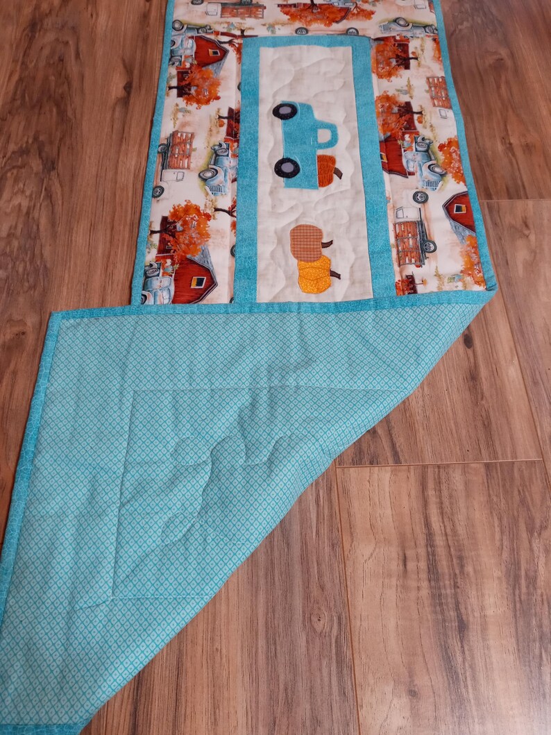 May include: A quilted table runner featuring a central panel with a blue truck and hay bales applique. The runner has a white background with a teal border and backing. The sides display a repeating pattern of red barns and fall foliage.