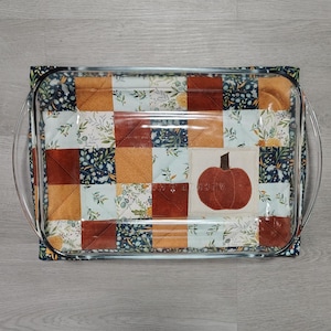 May include: A clear glass baking dish with a quilted, patchwork-style cozy. The cozy features squares of orange, burgundy, and floral patterned fabric. A central square displays a brown pumpkin. The dish has curved handles.