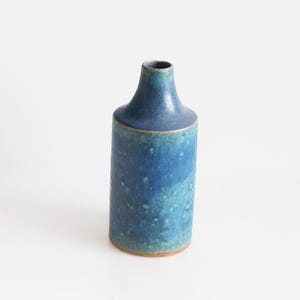 May include: A handcrafted ceramic vase with a cylindrical body and a narrow neck. The vase features a gradient of blue hues, from a deep indigo to a lighter turquoise, with a textured surface. The base is a natural, unglazed clay color.