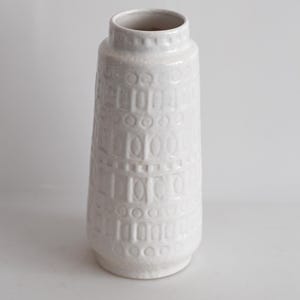 May include: A white ceramic vase with a textured surface. The vase has a cylindrical shape with a slightly flared base and a narrow opening. The surface is decorated with a repeating pattern of geometric shapes.