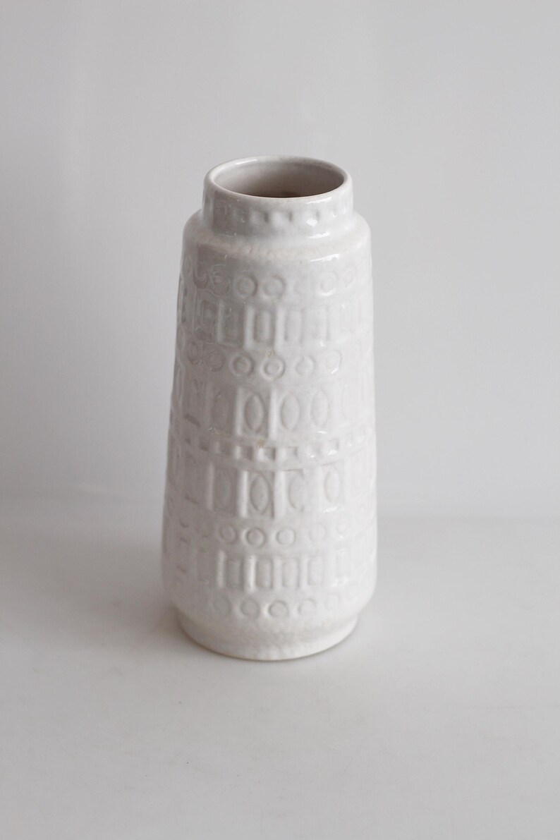 May include: A white ceramic vase with a textured surface. The vase has a cylindrical shape with a slightly flared base and a narrow opening. The surface is decorated with a repeating pattern of geometric shapes.