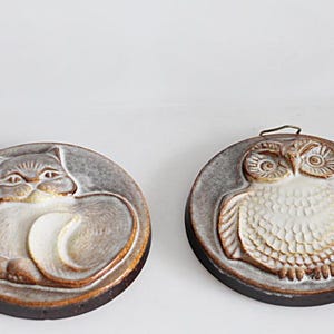 May include: Two ceramic wall plaques featuring animal designs. One depicts a cat in a resting pose, the other an owl. Both are circular with a gray background and brown trim. The owl plaque has a metal loop for hanging.