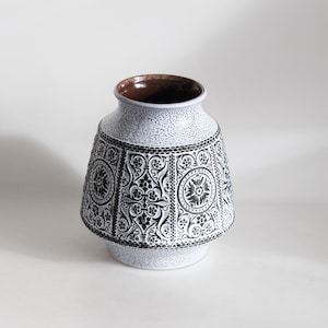 Op de afbeelding: Een witte keramische vaas met een getextureerd oppervlak en een uitlopende basis. De vaas heeft een zwart-wit bloemmotief rond de body en een bruin interieur. De vaas is een decoratief item.