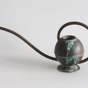Puede incluir: Una regadera esférica de cobre con un pico largo y delgado y un asa circular. La superficie metálica muestra una pátina marrón desgastada con parches visibles de cardenillo verde oxidado, apoyada sobre una pequeña base de pedestal acampanada.