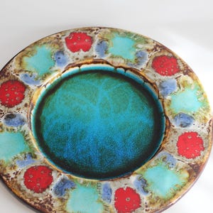Puede incluir: Un plato redondo de cerámica con un diseño vibrante. El plato presenta un área central turquesa rodeada por un anillo de acentos rojos, azules y turquesas. El borde tiene una apariencia marrón texturizada.