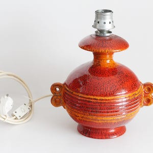 Könnte beinhalten: Ein Keramik-Lampenfuß mit einer leuchtend orange-roten, gesprenkelten Glasur und horizontalen Rillen in der Mitte. Die Lampe verfügt über zwei seitliche Griffe, einen ausgestellten oberen Bereich und ein weißes Stromkabel mit Stecker.