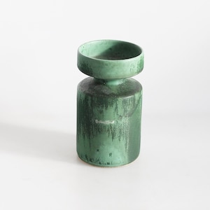 Könnte beinhalten: Eine grüne Keramikvase mit zylindrischem Fuß und rundem Oberteil. Die Vase hat eine strukturierte Oberfläche mit dunkelgrünen Streifen. Die Oberseite ist ein hellerer Grünton. Die Vase ist etwa 18 cm hoch.