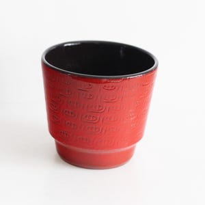 Op de afbeelding: Een rode keramische bloempot met een zwart interieur. De buitenkant heeft een herhalend reliëfpatroon. De pot heeft een licht taps toelopende vorm en een afgeronde basis. Geschikt voor kleine planten.