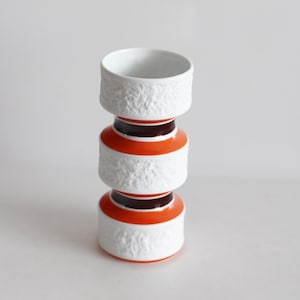 Könnte beinhalten: Eine Vase aus Keramik in Weiß und Orange. Die Vase hat eine strukturierte Oberfläche und eine zylindrische Form mit drei gestapelten Abschnitten. Die oberen und mittleren Abschnitte sind weiß mit einem strukturierten Muster, während die mittleren und unteren Abschnitte orange sind.