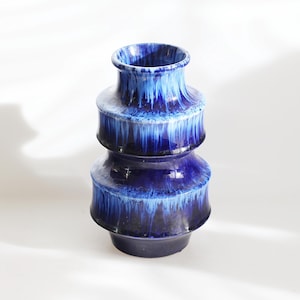 Puede incluir: Un jarrón de cerámica con un diseño en capas y un acabado brillante. El jarrón presenta un degradado de azules, que va desde un índigo profundo en la base hasta tonos más claros en la parte superior. El jarrón mide unos 25 cm de altura.