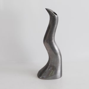 May include: A silver metal vase with a unique, abstract design. The vase is tall and slender, with a curved shape that resembles a bird's neck.