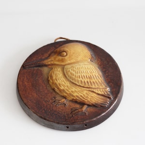 Op de afbeelding: Ronde, bruine keramische wanddecoratie met een gedetailleerd reliëf van een vogel. De vogel is afgebeeld in tinten bruin en beige, met ingewikkelde veerdetaillering. Het heeft een lus om op te hangen.