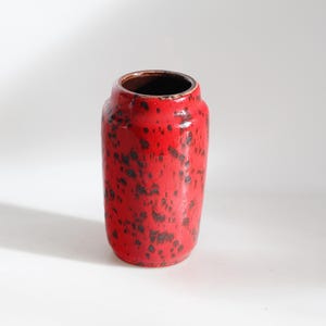 Puede incluir: Un jarrón cilíndrico de cerámica roja con un patrón moteado de manchas marrón oscuro. El jarrón tiene una abertura ancha y un interior marrón oscuro. El jarrón mide aproximadamente 20 cm de alto.