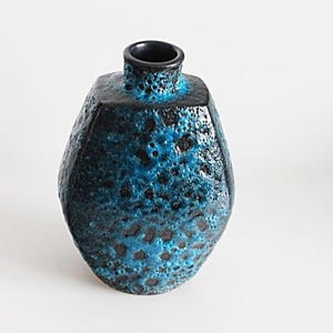 May include: A ceramic vase with a textured surface, featuring a dark blue and turquoise glaze. The vase has a distinctive shape with a narrow neck and a wider body. The surface is speckled with dark spots. A decorative item for the home.
