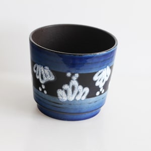 Op de afbeelding: Een keramische plantenbak met een donkerblauwe binnen- en buitenkant. De buitenkant heeft een zwarte band met witte bloemmotieven, geplaatst tussen banden van lichter blauw. De plantenbak is cilindrisch met een licht schuine rand.