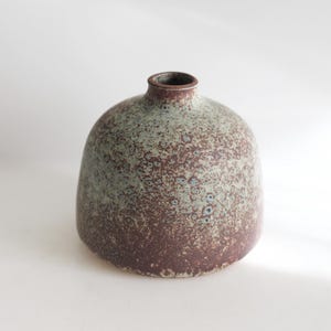 May include: A small, speckled ceramic vase with a rounded body and a narrow neck. The vase features a color gradient, transitioning from a deep burgundy at the base to a muted green near the top. The surface has a textured, speckled appearance.