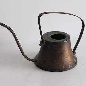 Könnte beinhalten: Eine kleine, antike Gießkanne aus Kupfer mit einem langen, gebogenen Ausguss und einem geschwungenen Griff. Die Kanne hat einen konischen Körper und eine dunkle, gealterte Patina, was auf einen Vintage-Zustand hindeutet.