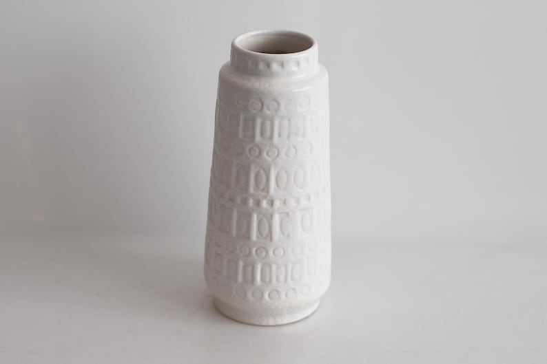 May include: A white ceramic vase with a textured, geometric pattern. The vase has a cylindrical shape with a slightly flared base and a narrow opening at the top. The design features repeating rectangular and circular shapes.