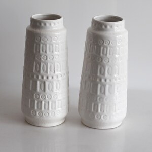 May include: Two white ceramic vases with a textured, geometric pattern. The vases have a cylindrical shape with a slightly flared base and a narrow neck. The surface is decorated with a repeating pattern of rectangles and circles.