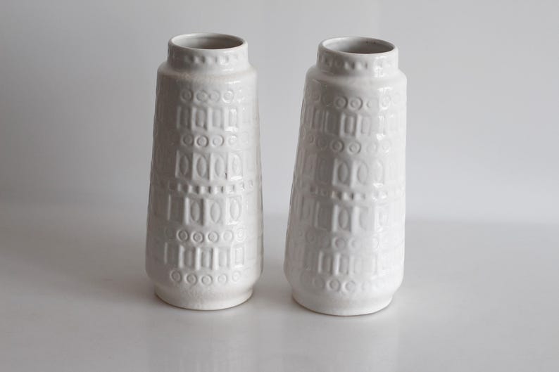 May include: Two white ceramic vases with a textured, geometric pattern. The vases have a cylindrical shape with a slightly flared base and a narrow neck. The surface is decorated with a repeating pattern of rectangles and circles.