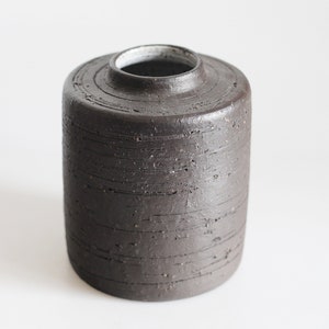 Könnte beinhalten: Eine dunkelbraune Keramikvase mit einer strukturierten, gestreiften Oberfläche. Die Vase hat eine breite, runde Öffnung.