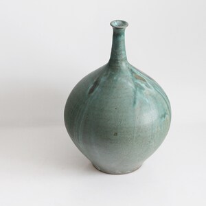 May include: A teal ceramic vase with a rounded body and a slender neck. The vase has a matte finish and subtle colour variations, adding to its unique, artistic appeal. This decorative piece is ideal for displaying flowers or as a standalone art object.