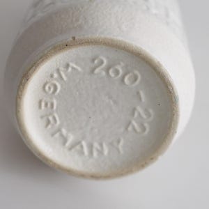 May include: Close-up of the bottom of a white ceramic vase. The base is circular with the embossed text "W.GERMANY 260-22". The vase has a textured surface and a slightly tapered shape. The overall aesthetic is minimalist and vintage.