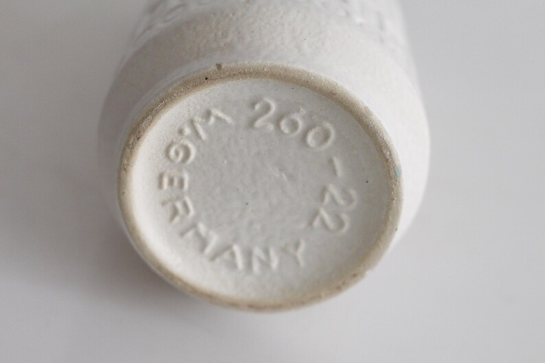 May include: Close-up of the bottom of a white ceramic vase. The base is circular with the embossed text "W.GERMANY 260-22". The vase has a textured surface and a slightly tapered shape. The overall aesthetic is minimalist and vintage.