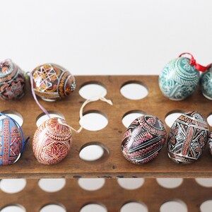 Può includere: Un porta uova in legno con una varietà di uova di Pasqua dipinte in modo intricato. Le uova sono decorate con motivi geometrici in tonalità di blu, verde, rosso e nero.