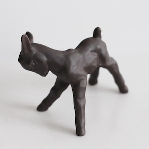 Puede incluir: Figura de cerámica marrón oscuro de un joven ciervo en una pose juguetona. La escultura tiene un acabado liso y está colocada sobre un fondo blanco liso. El ciervo está representado con la cabeza baja y las patas en posición de caminar.