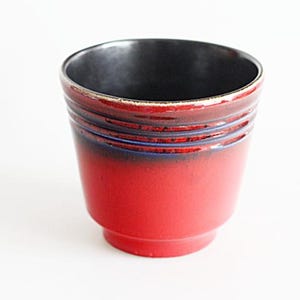 Könnte beinhalten: Ein roter Keramiktopf mit schwarzem Innenraum. Der Topf hat eine leicht ausgestellte Form mit horizontalen Rillen und einem dunkelblauen Band in der Nähe der Oberseite. Die Außenfarbe geht von Rot in Schwarz über.
