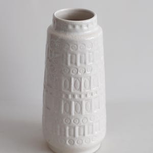 May include: A white ceramic vase with a textured surface. The vase has a cylindrical shape with a slightly flared base and a narrow neck. The surface is decorated with a repeating pattern of rectangles and circles.