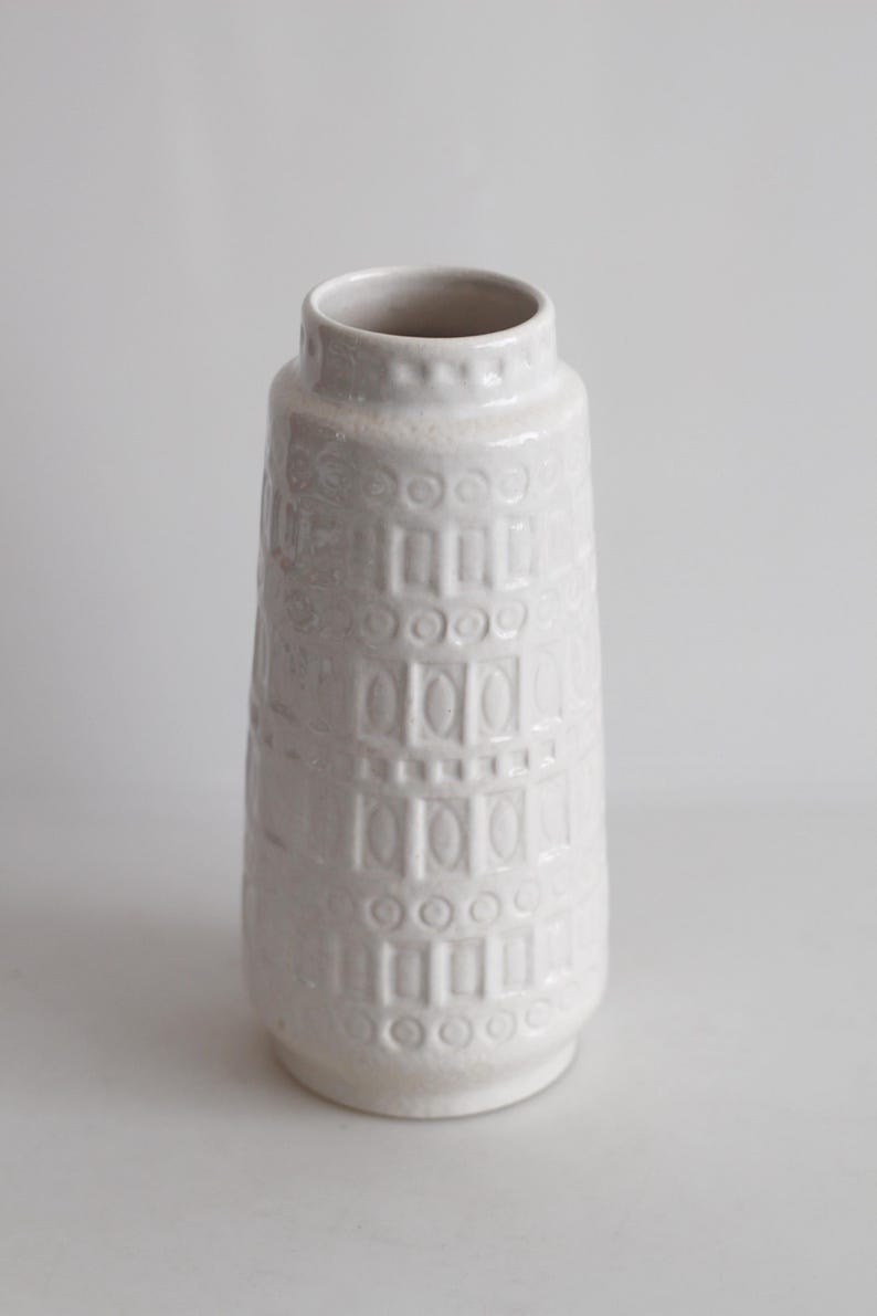 May include: A white ceramic vase with a textured surface. The vase has a cylindrical shape with a slightly flared base and a narrow neck. The surface is decorated with a repeating pattern of rectangles and circles.