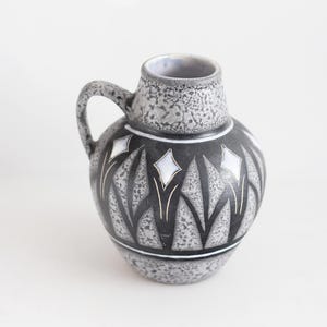 Puede incluir: Jarra de cerámica vintage con asa, con acabado gris moteado en la parte superior e inferior. El cuerpo principal es negro con un diseño de diamantes y hojas blancas. La jarra tiene forma redondeada y una pequeña abertura.