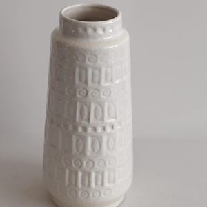 May include: A white ceramic vase with a textured surface. The vase has a cylindrical shape with a slightly flared base and a narrow opening. The surface is decorated with a repeating pattern of rectangles and circles.