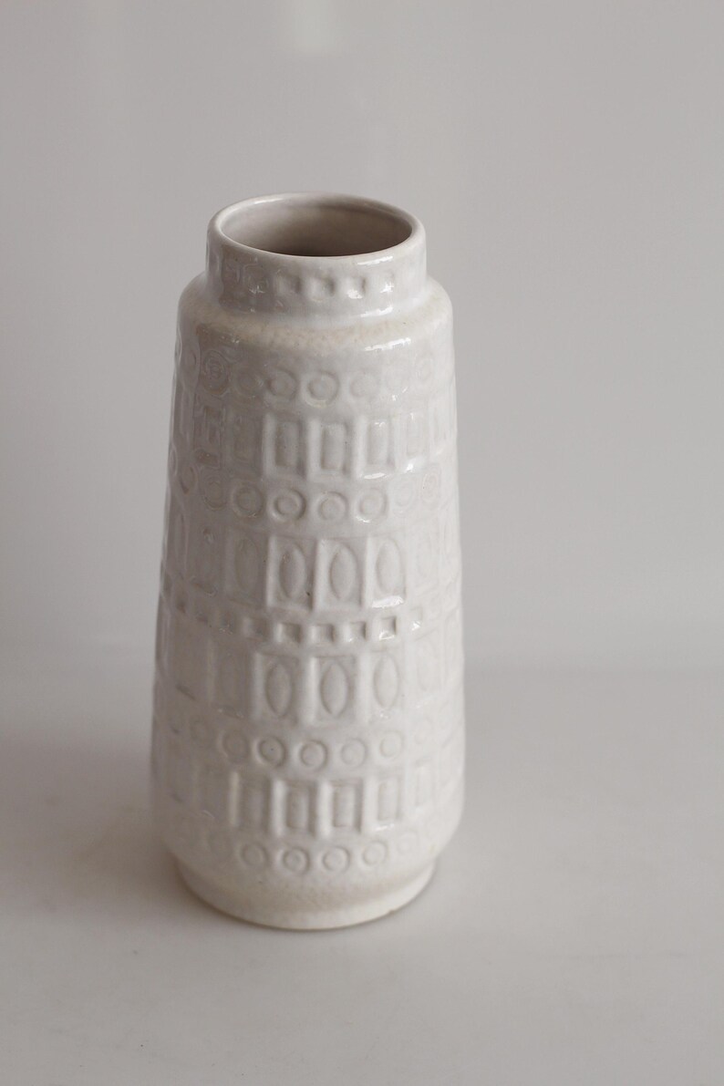 May include: A white ceramic vase with a textured surface. The vase has a cylindrical shape with a slightly flared base and a narrow opening. The surface is decorated with a repeating pattern of rectangles and circles.