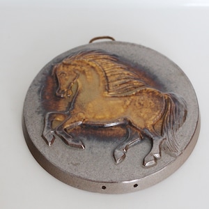 Op de afbeelding: Een keramische wandhanger met een bruin paard in reliëf, tegen een grijze achtergrond. Het paard is afgebeeld in een rennende pose met een stromende manen. Het stuk heeft een ronde vorm en een rustieke afwerking.