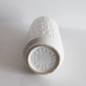 May include: A white ceramic vase with a textured, geometric pattern. The vase is cylindrical with a slightly flared base. The bottom of the vase has a circular stamp with text and the number 260.