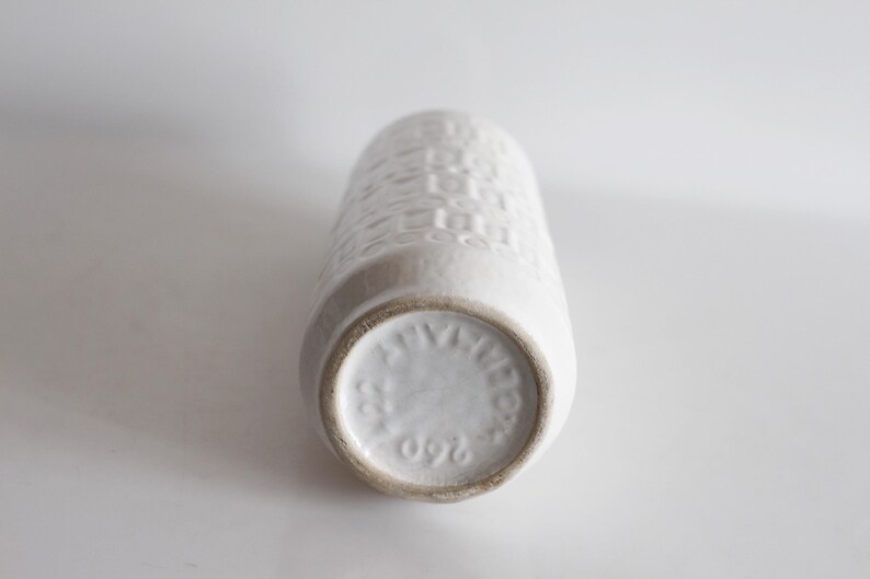 May include: A white ceramic vase with a textured, geometric pattern. The vase is cylindrical with a slightly flared base. The bottom of the vase has a circular stamp with text and the number 260.