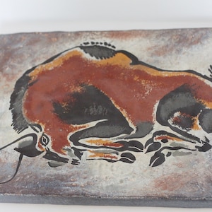 Puede incluir: Un azulejo de cerámica con una imagen estilizada de un bisonte marrón y negro, pintado en estilo de pintura rupestre.