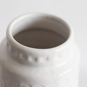 May include: Close-up of a white ceramic jar with a wide opening. The jar has a slightly textured surface and a series of small, decorative indentations around the upper portion. The interior is a darker shade, creating contrast.