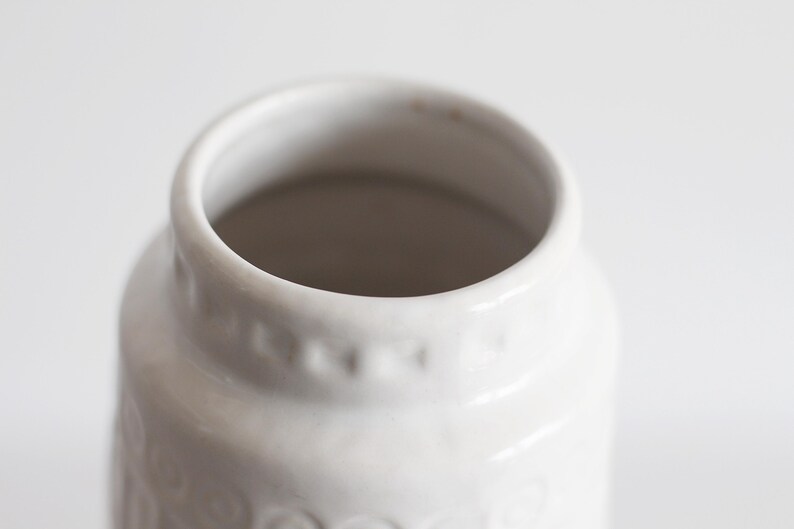 May include: Close-up of a white ceramic jar with a wide opening. The jar has a slightly textured surface and a series of small, decorative indentations around the upper portion. The interior is a darker shade, creating contrast.