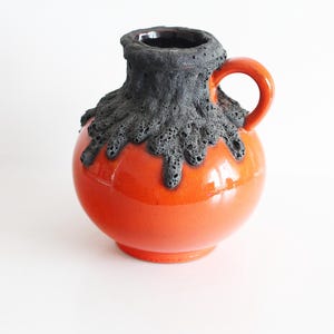 Puede incluir: Un jarrón de cerámica con un cuerpo redondo naranja y una parte superior negra texturizada. El jarrón tiene un pequeño asa naranja y un acabado brillante, adecuado para la decoración del hogar.