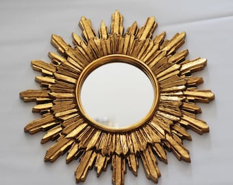May include: A gold sunburst mirror with a round, reflective surface. The mirror has a decorative, ornate frame with a sunburst design.